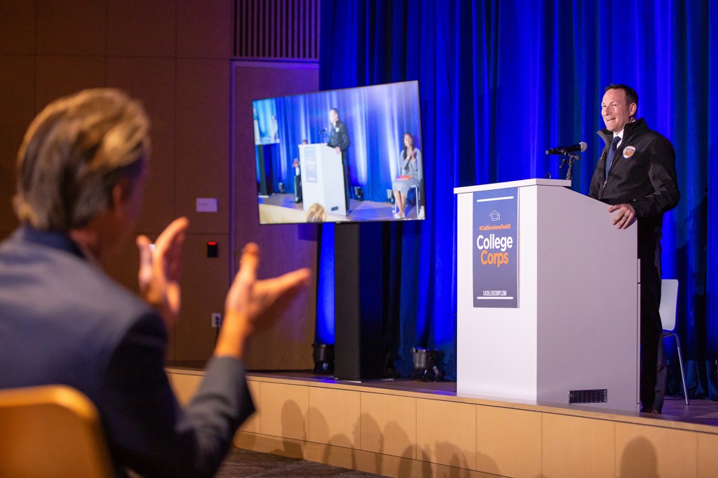 A speaker stands at a podium labeled “College Corps,” addressing an audience while on a stage. On the left side of the frame is a person out of focus, clapping while they listen to the speaker on the stage.