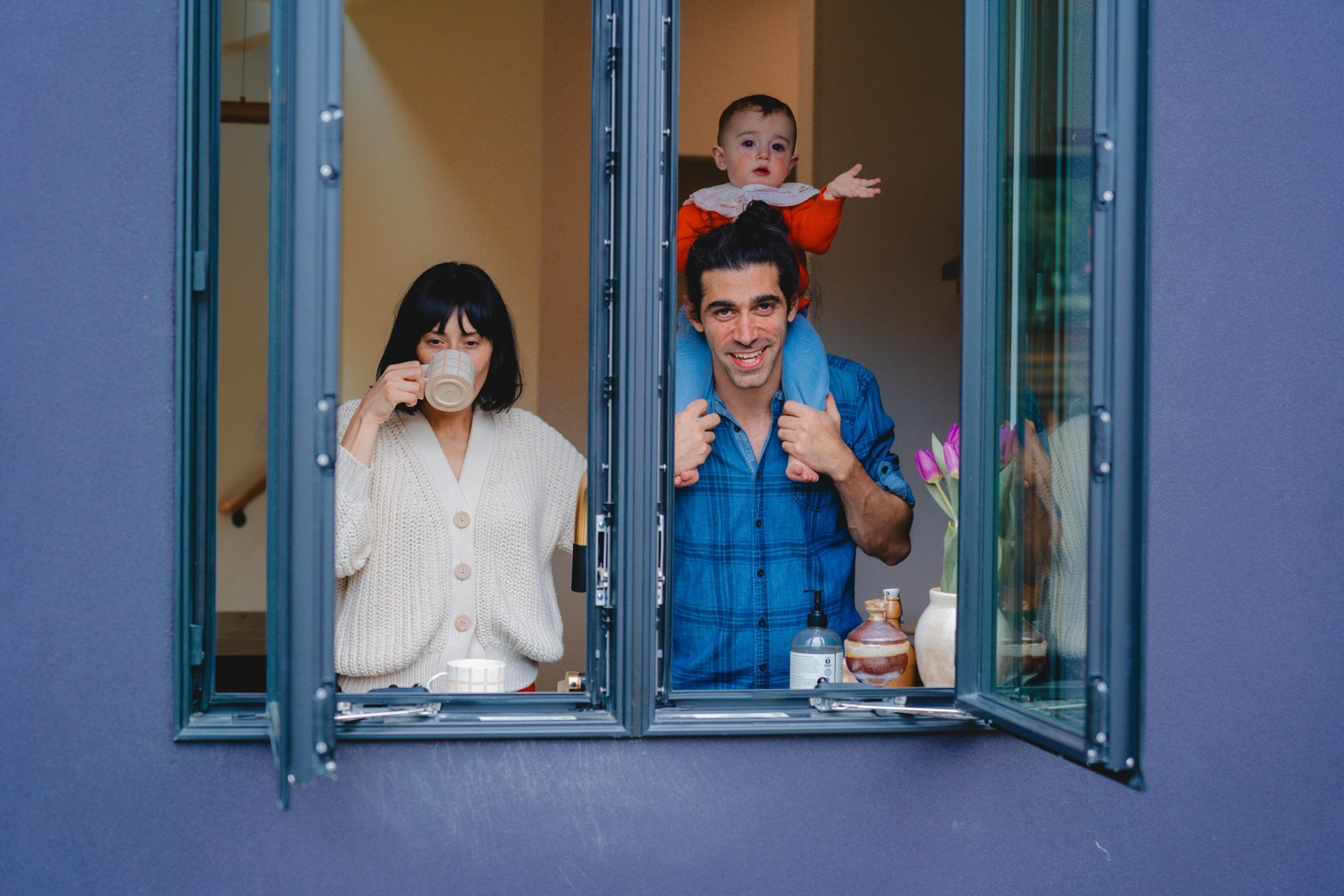 A man wearing a blue shirt carries a baby on his shoulders while a woman in a cream sweater drinks from a mug, both standing by an open window.