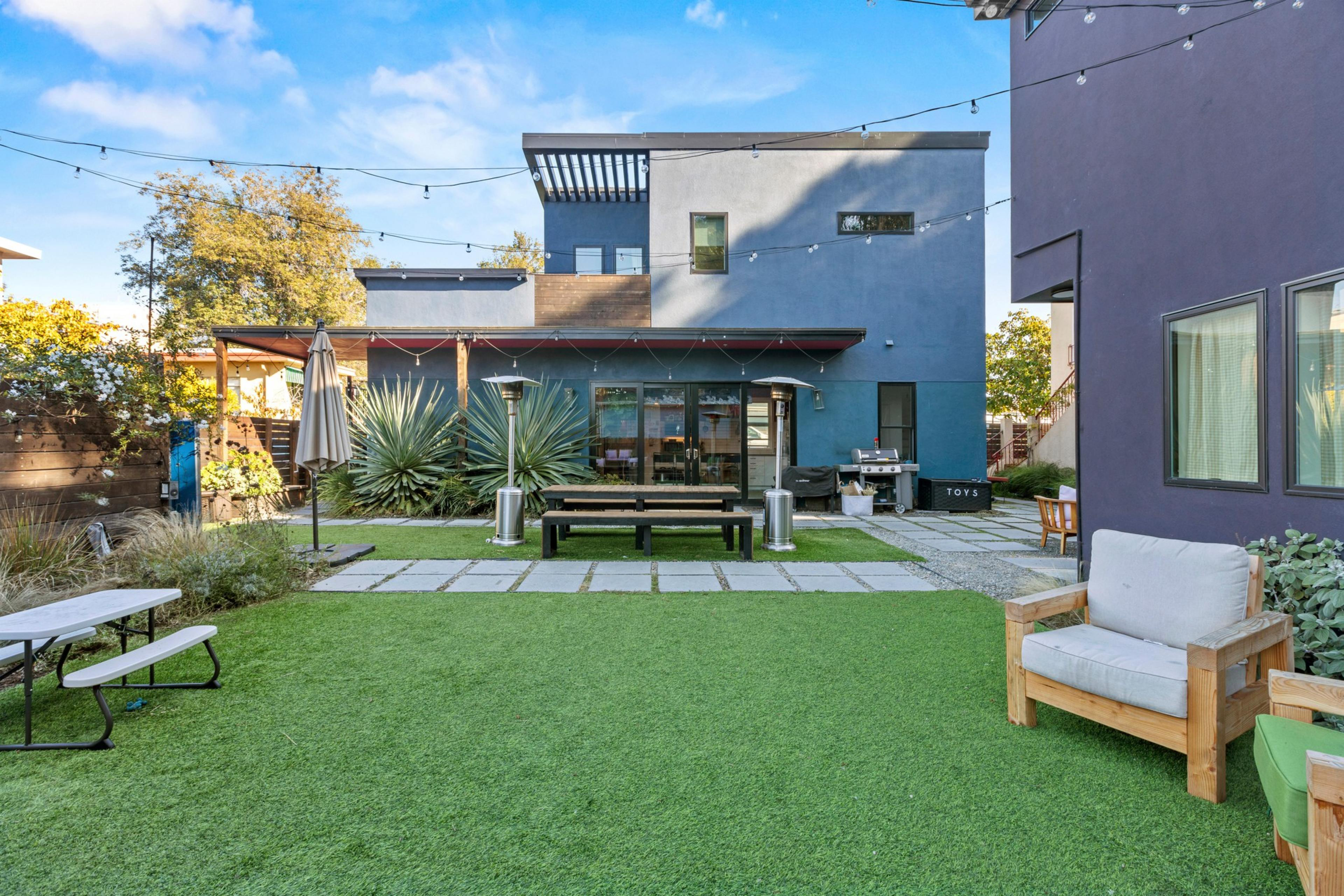 A modern backyard with artificial grass, a picnic table, wooden bench, outdoor heater, umbrella, and blue-gray two-story house under a blue sky.