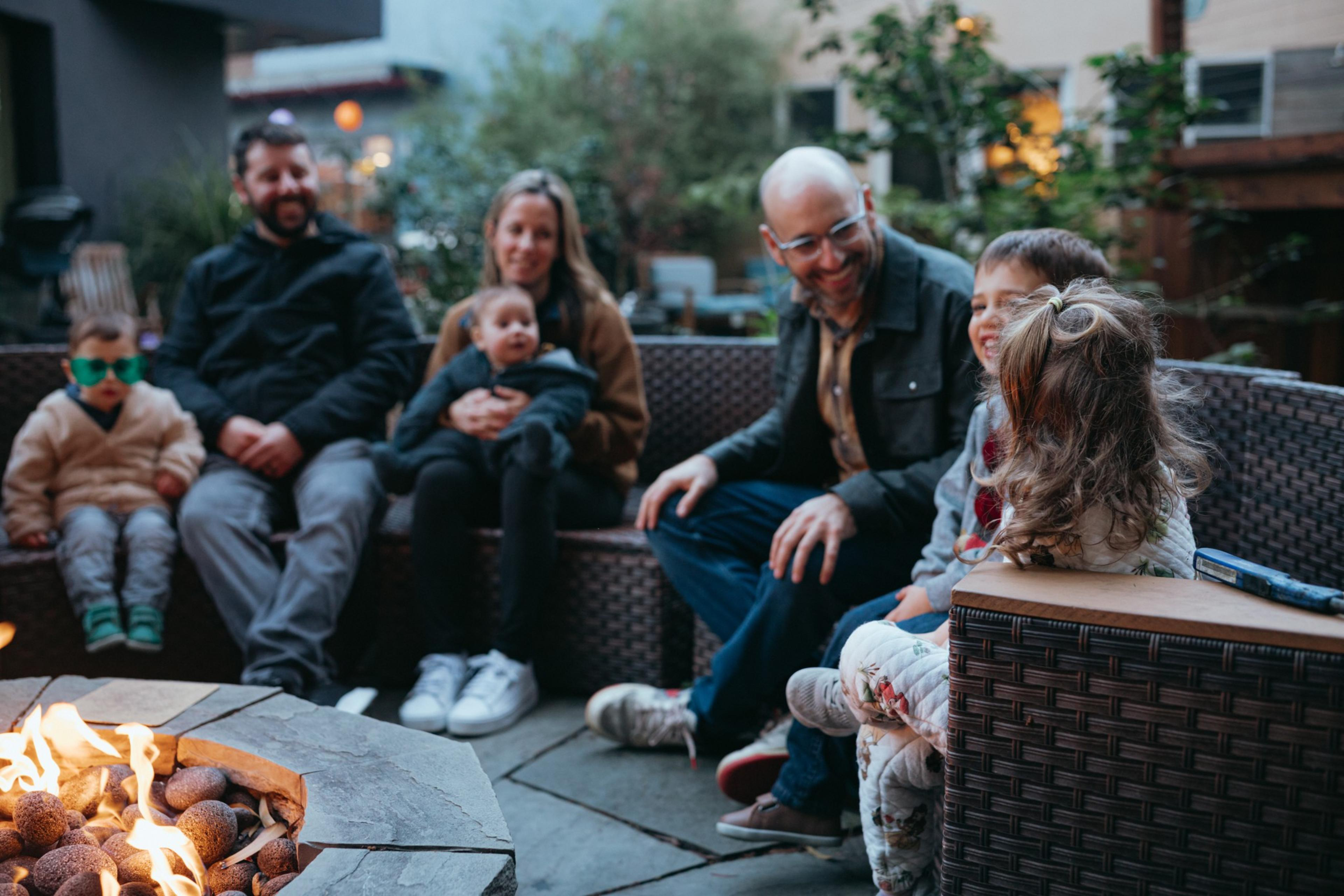 Six people, including three children and three adults, sit and smile around a fire pit outside in a cozy backyard setting.