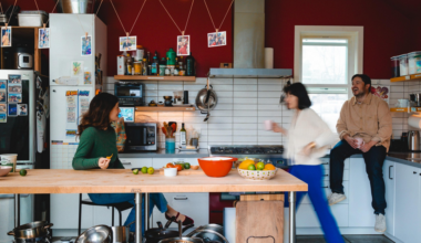Three people in a kitchen: one seated at a counter eating, one walking with a cup, and one sitting on the countertop laughing, with colorful fruits on the table.