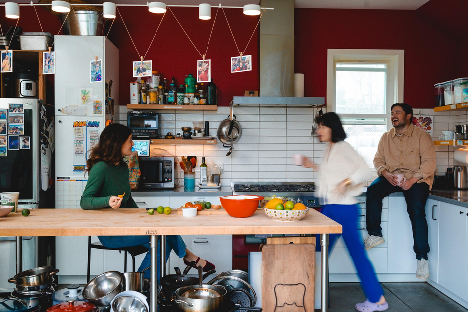 Three people in a kitchen: one seated at a counter eating, one walking with a cup, and one sitting on the countertop laughing, with colorful fruits on the table.