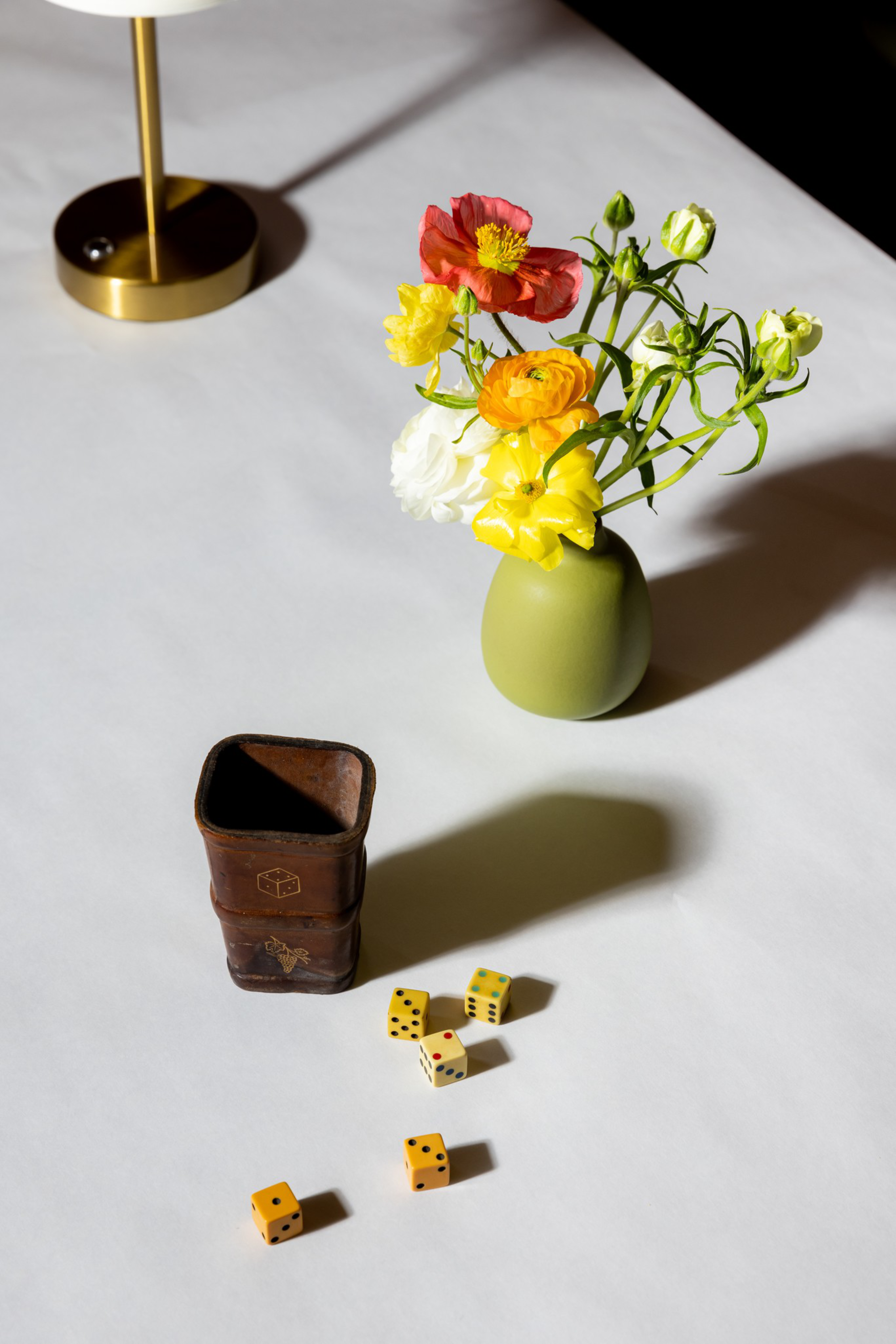 A green vase with colorful flowers, a brass lamp base, a small brown leather dice cup, and six yellow dice scattered on a white tablecloth.