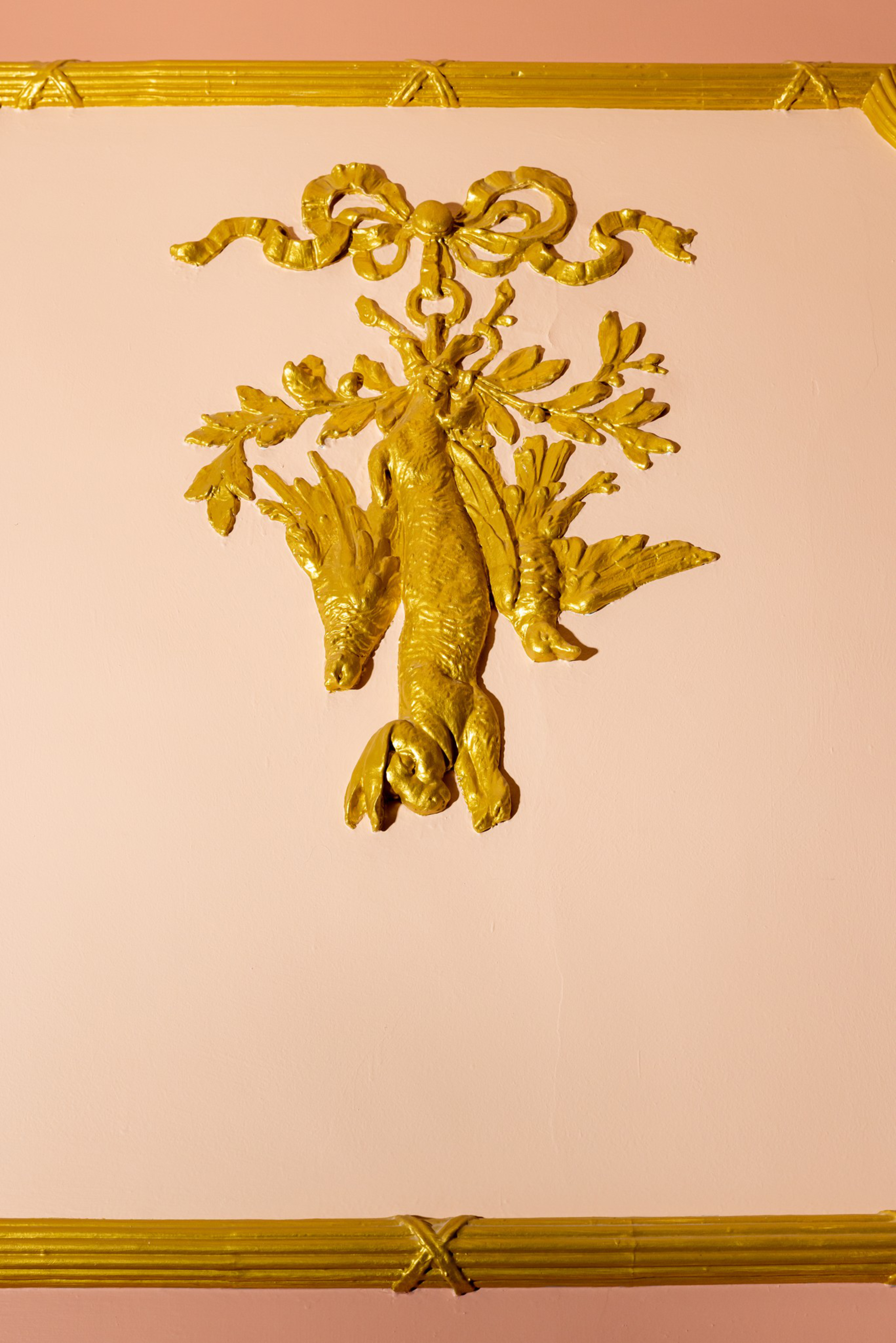 A golden wall decoration features a hanging rabbit and two birds, surrounded by leaves and topped with a decorative bow, against a pale pink background.