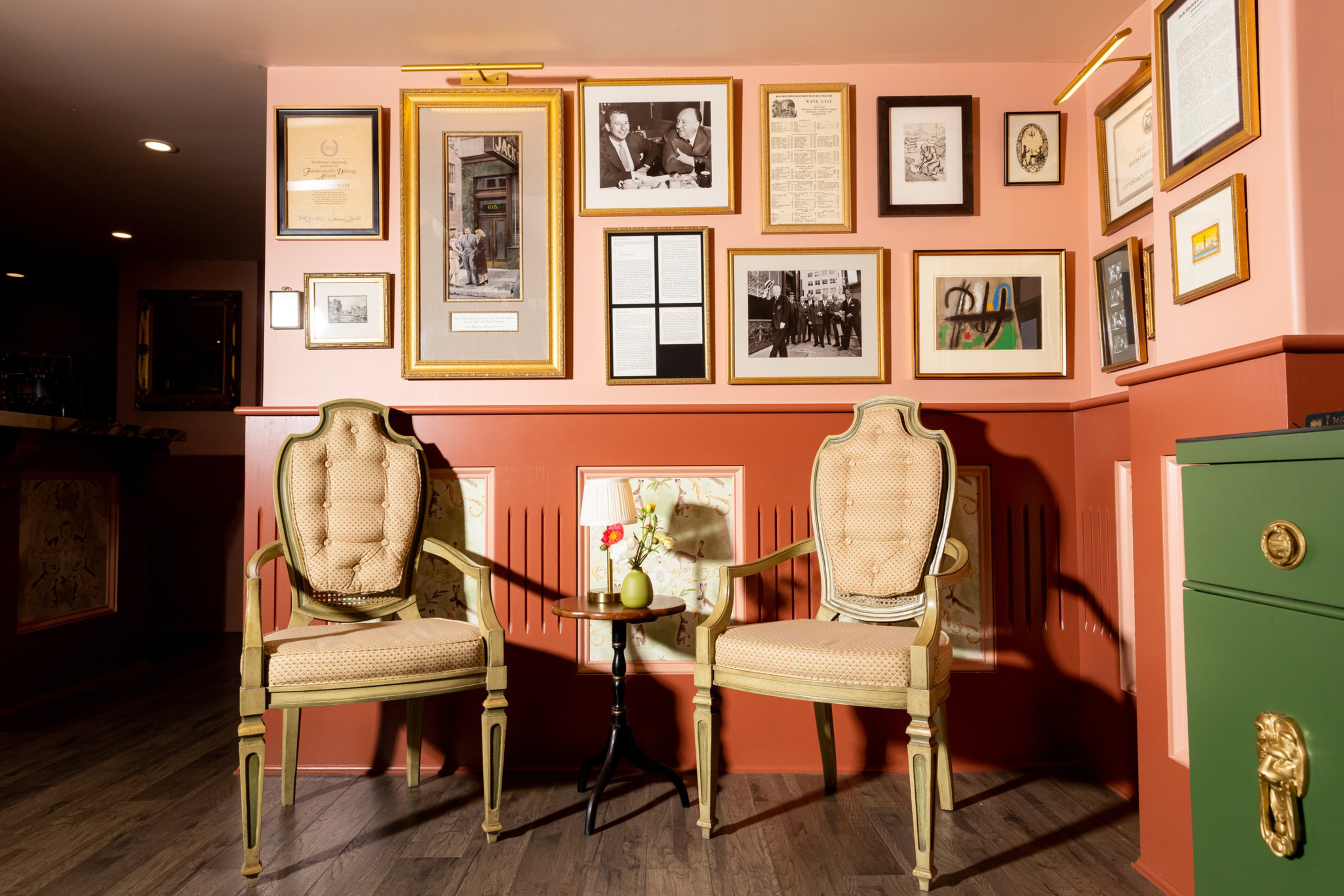 Two vintage upholstered chairs flank a small round table with a lamp and flower, set against a wall adorned with various framed pictures and documents.