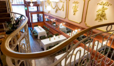 A cozy dining area with white tablecloths, green chairs, ornate gold wall decorations, and a curved white railing overlooking the space.
