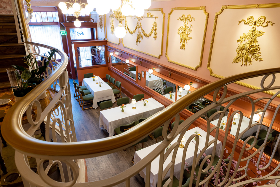 A cozy dining area with white tablecloths, green chairs, ornate gold wall decorations, and a curved white railing overlooking the space.