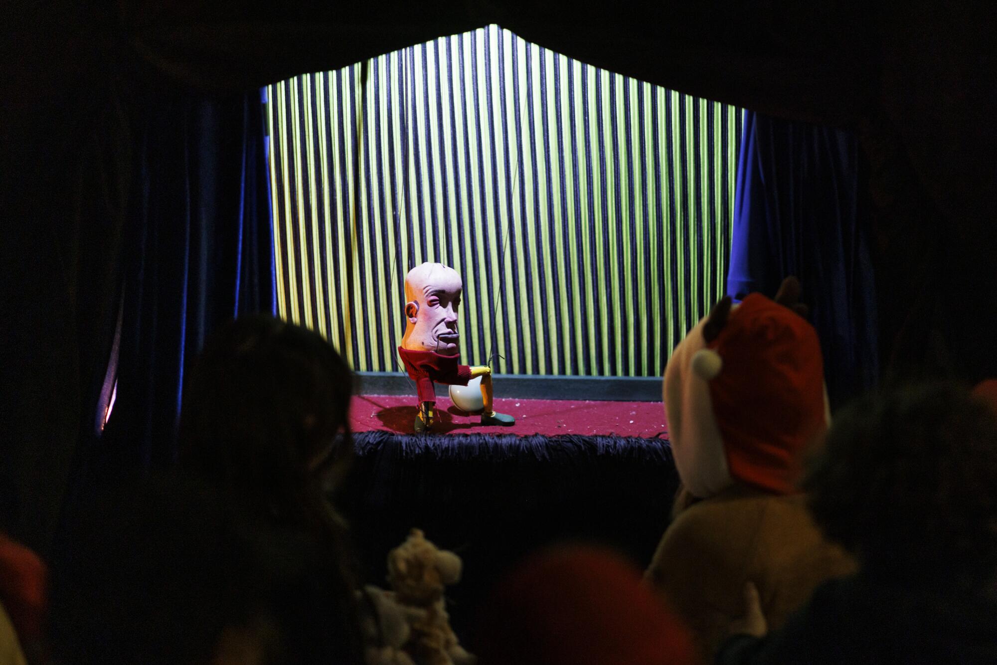 A puppet with a huge egg head on a body of red pants.