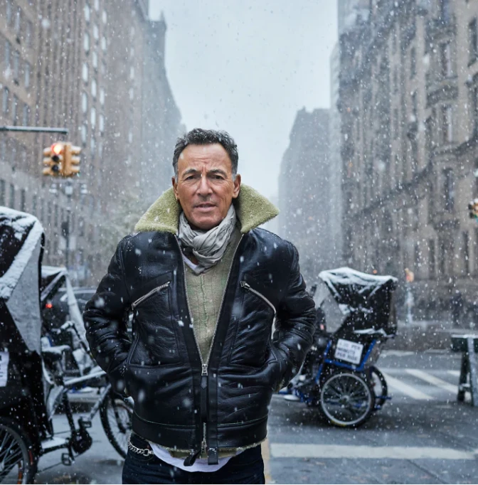 A white man in a black leather jacket stands in the street while it snows.