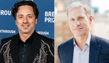A man with dark wavy hair wears a black jacket with silver embroidery, smiling in front of a blue "Breakthrough Prize" backdrop; another man with short grey hair wears a blue checkered suit and smiles softly against a blurred cityscape background.