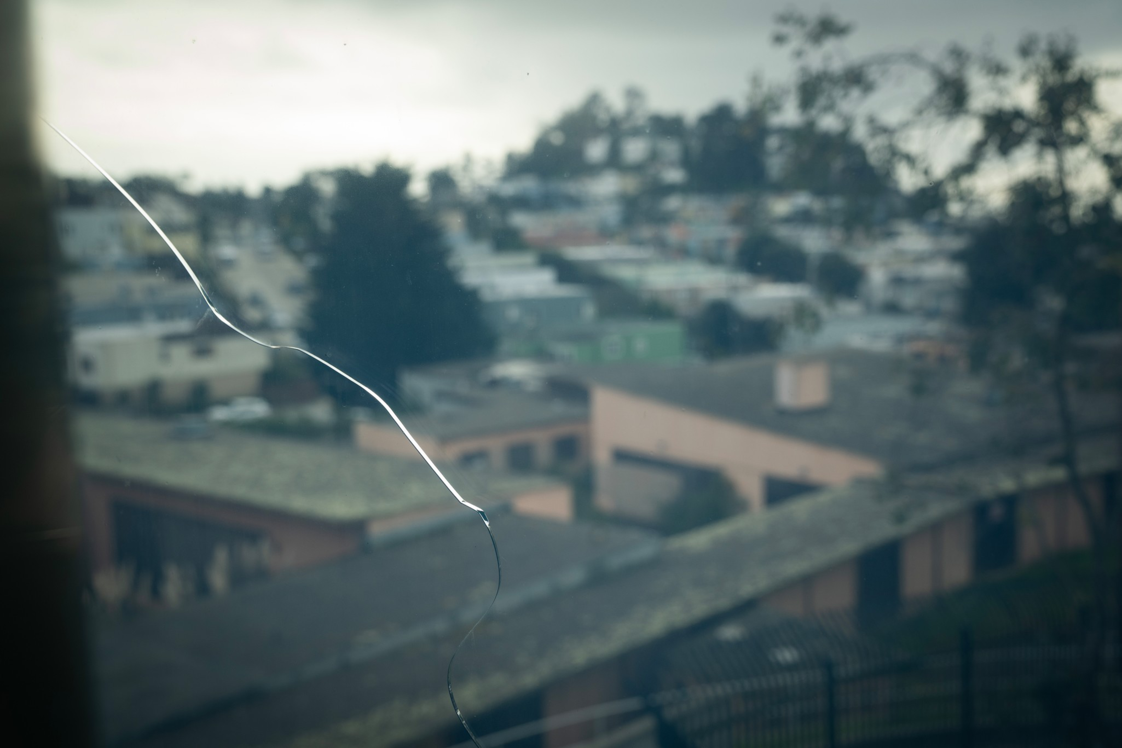 A cracked window pane with a faint outdoor view of buildings, trees, and a cloudy sky in the background.