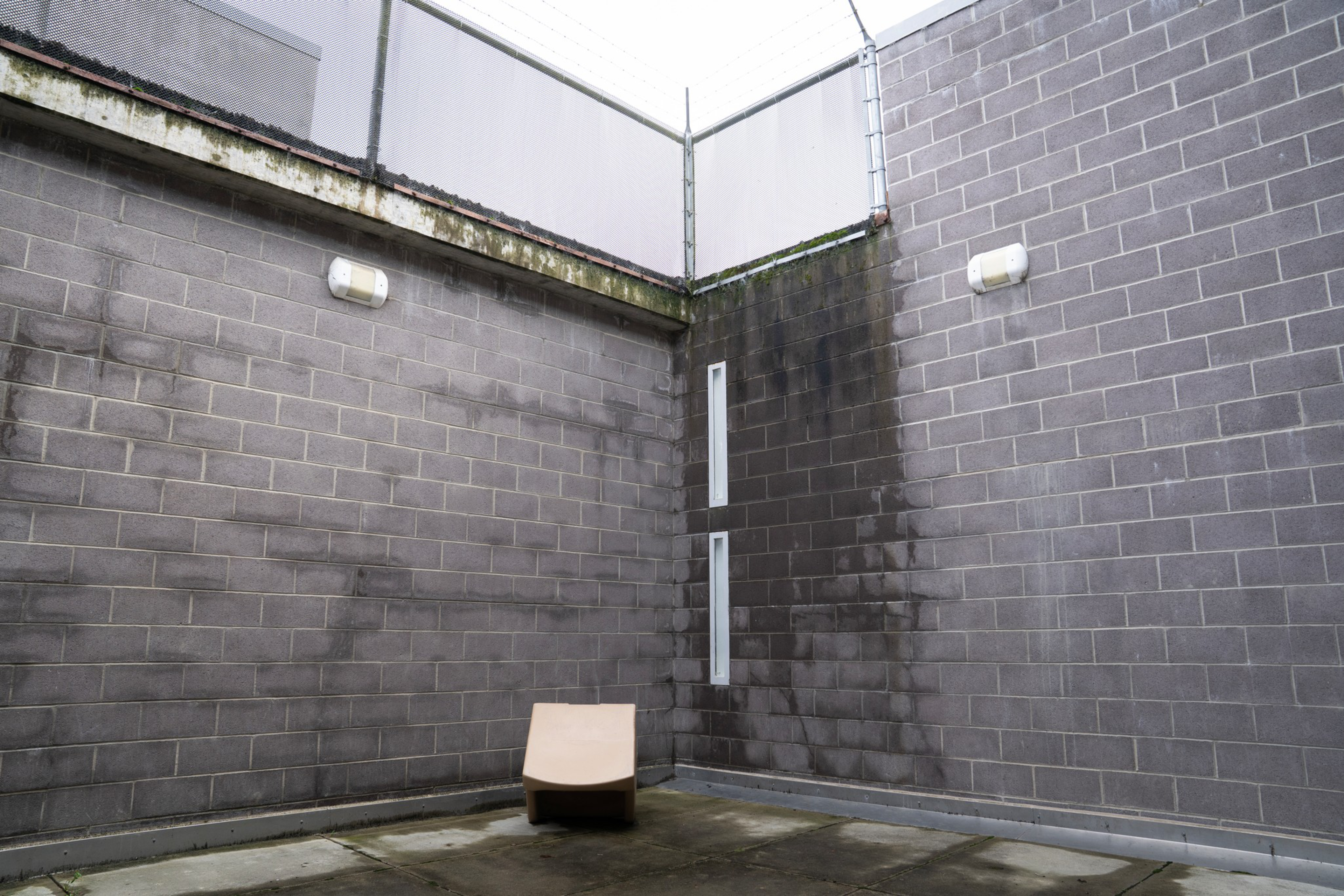 A gray concrete-walled enclosed outdoor space with a single beige plastic chair and a metal fence on top of the walls.