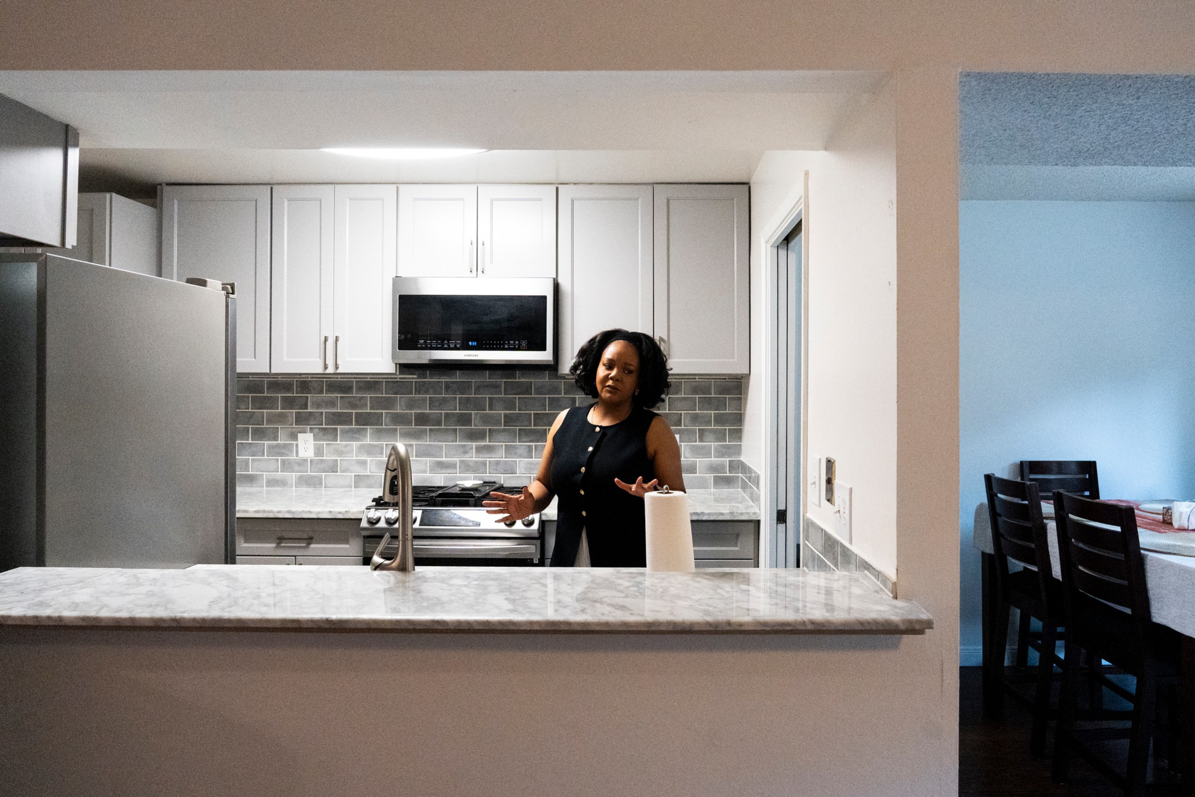 A woman in a black sleeveless top stands behind a kitchen counter with a paper towel roll, gesturing with her hands in a modern kitchen.