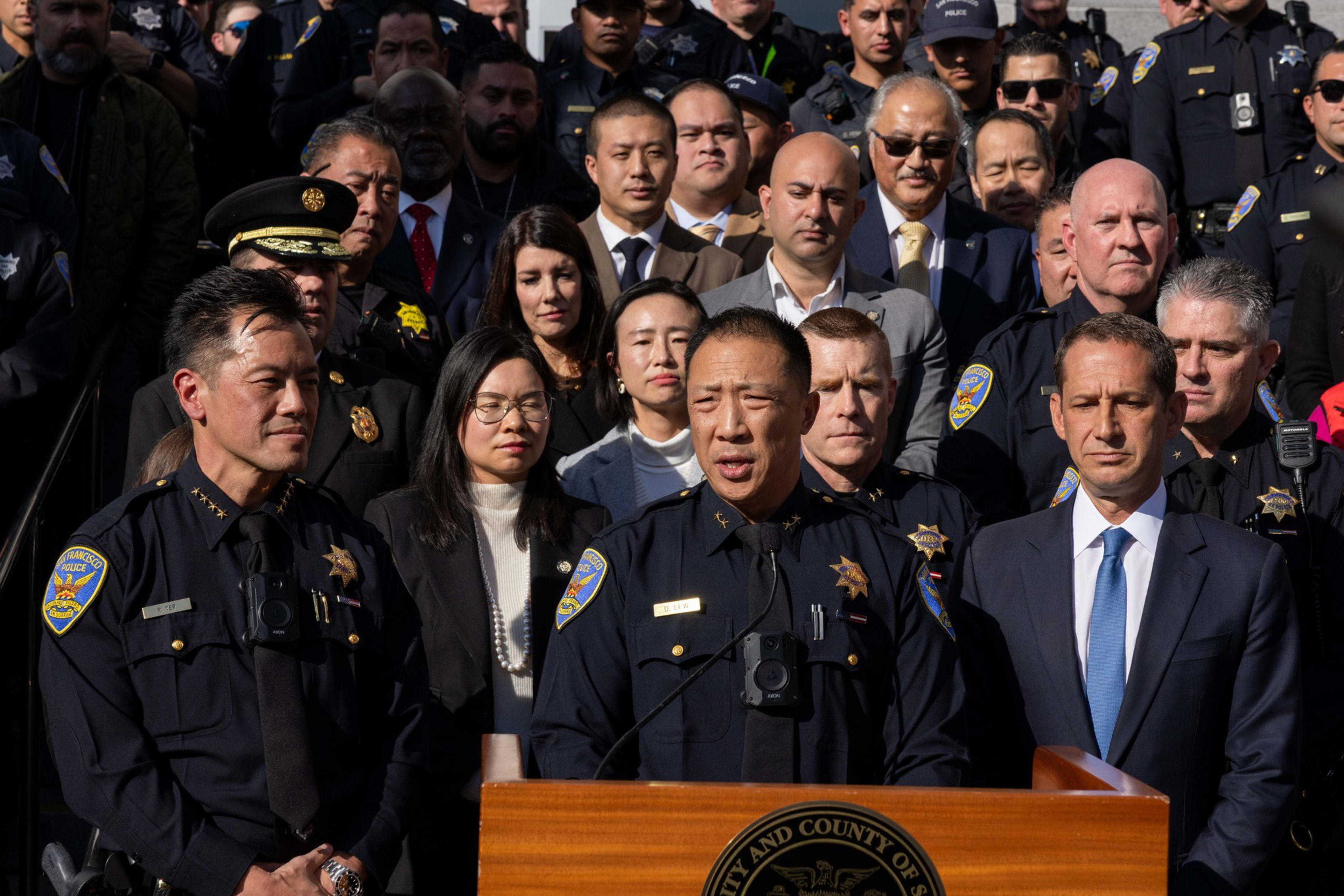 A group of police officers and officials stand closely together, with one officer speaking at a wooden podium bearing an official seal.