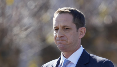 A man in a dark suit and light blue tie gazes pensively, set against a blurred outdoor background with soft lighting.