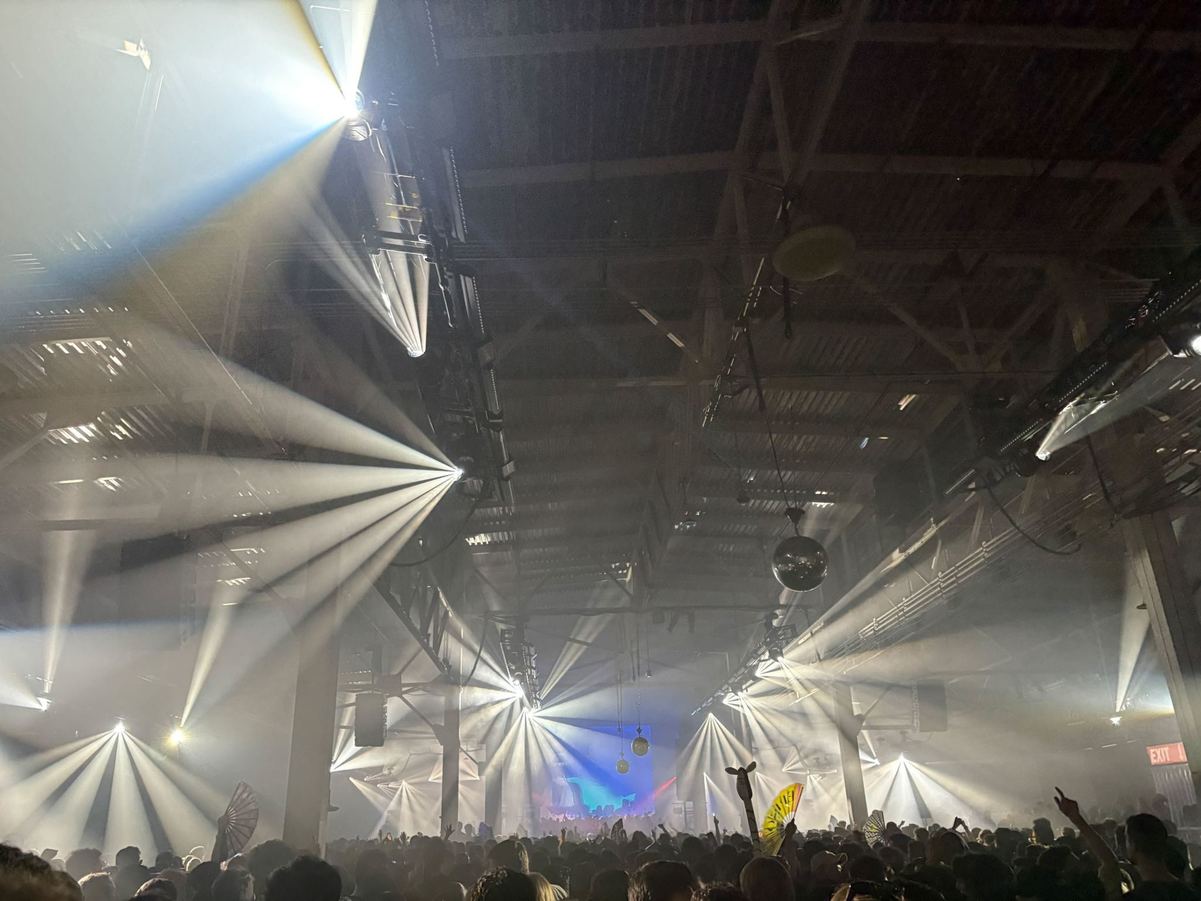 Bright stage lights beam through a foggy warehouse ceiling over a crowded concert with raised hands and a visible disco ball.