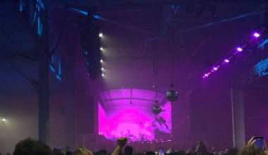 A large crowd watches a stage with bright purple lighting and projections in a dimly lit indoor venue with disco balls hanging from the ceiling.