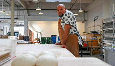 A shaven-headed man wearing a patterned short-sleeve shirt and a khaki apron stands next to a flat white table. Round balls of sourdough sit within his reach.