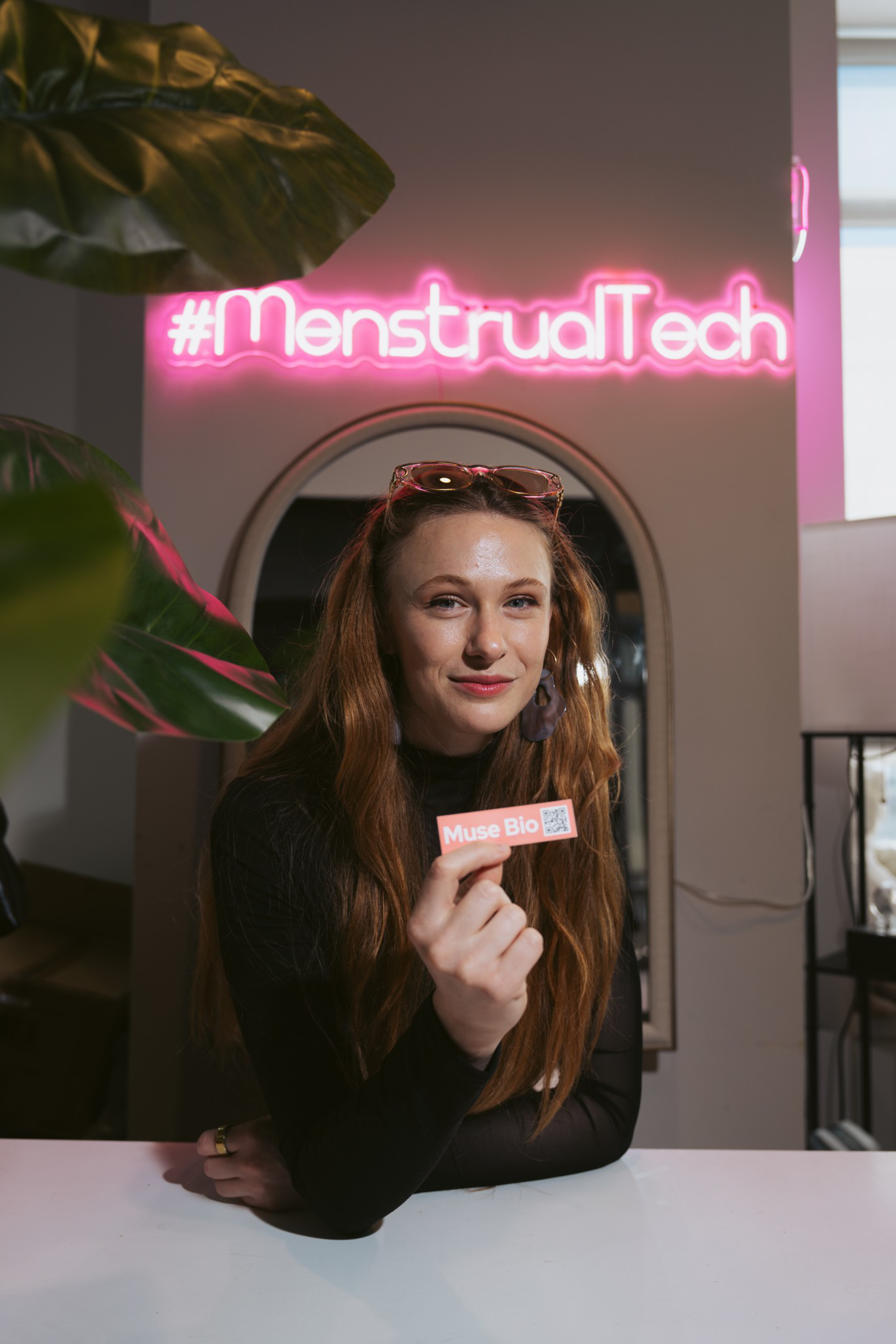 A woman with long brown hair holds a “Muse Bio” card, standing behind a white counter under a bright pink neon sign that reads “#MenstrualTech.”