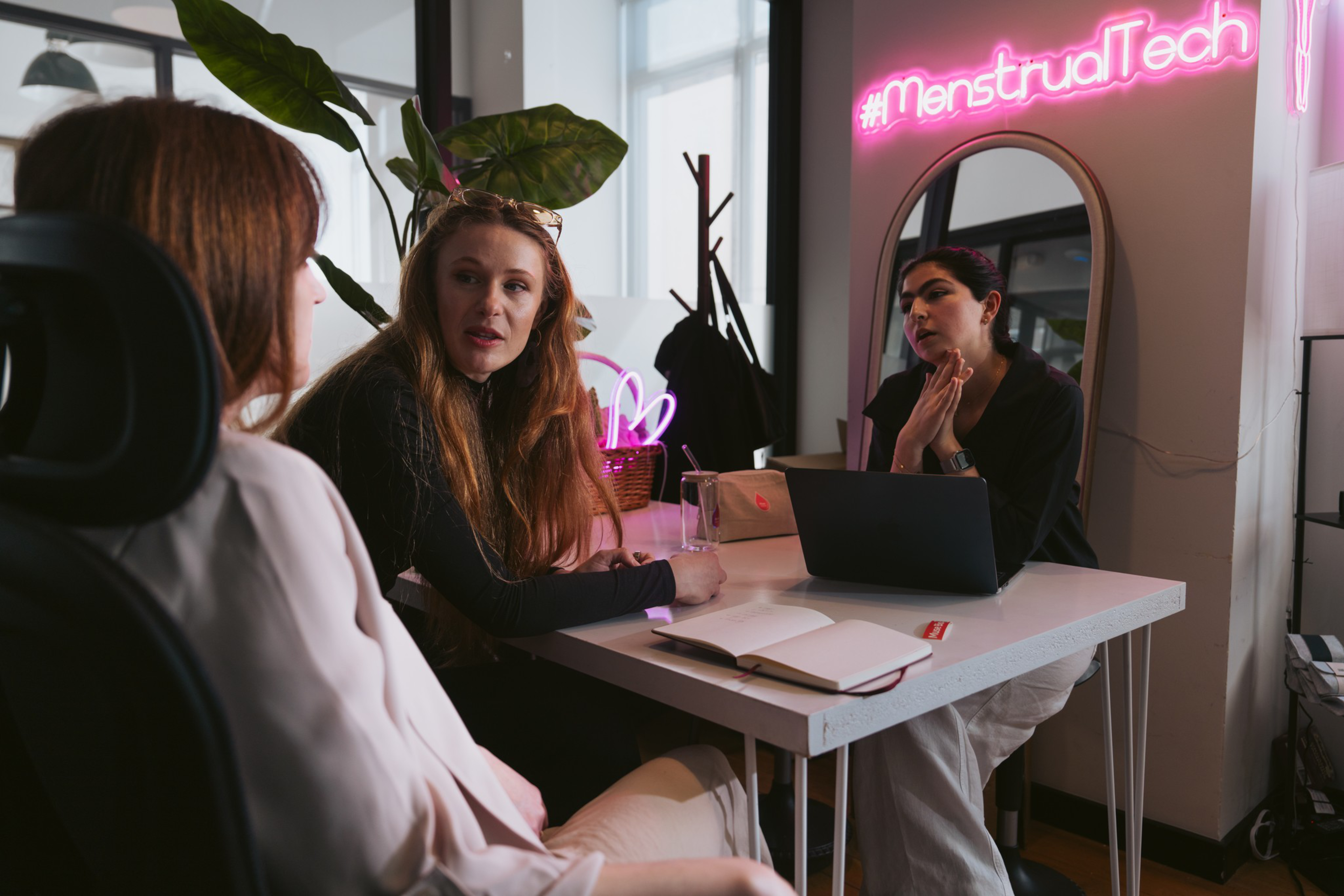 Three women are having a discussion around a white table with a laptop, notebook, and a glass in a room lit by a neon sign that reads “#MenstrualTech.”