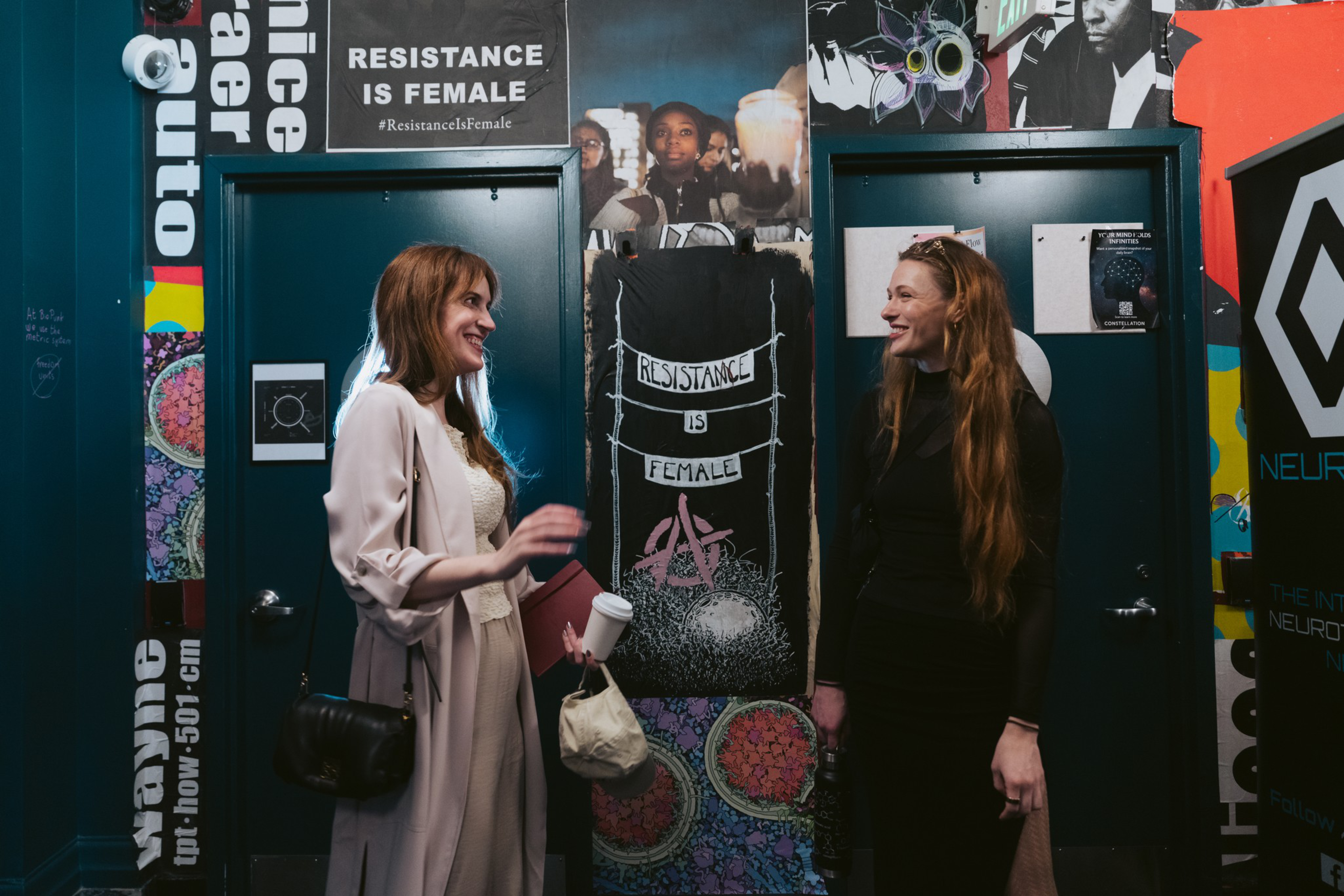 Two women are talking and smiling in front of a wall covered with colorful posters and graffiti, one of which reads “RESISTANCE IS FEMALE.”