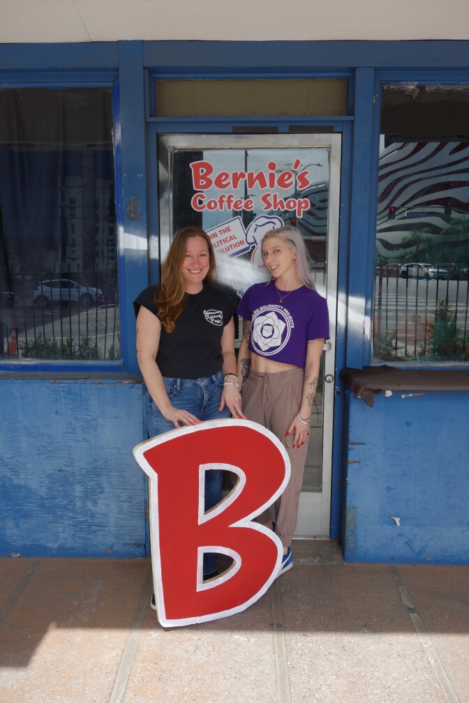 Two women stand outside a sign that reads "Bernie's Coffee Shop" with a large "B" sign.