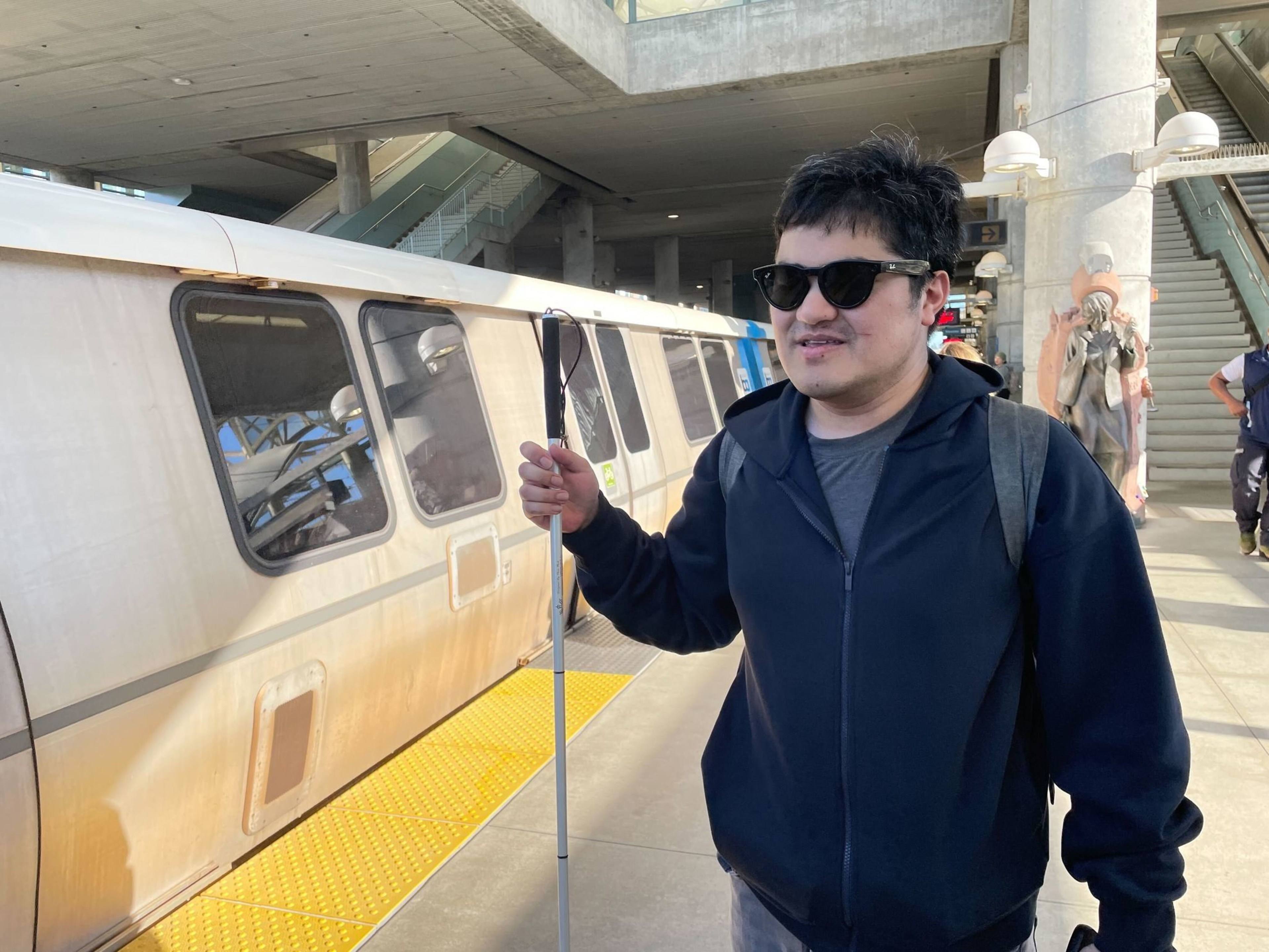 A man wearing sunglasses and a dark jacket holds a white cane near a train platform with a train and a stairway in the background.