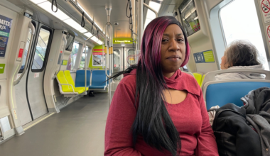 A woman with long black and pink hair wearing a red top sits on a mostly empty train with colorful blue and yellow seats.
