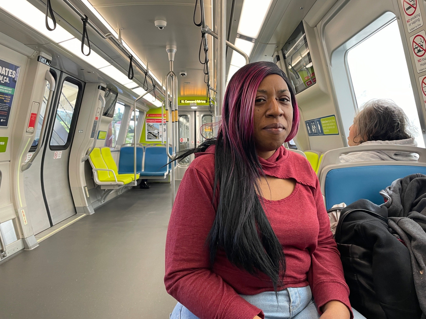 A woman with long black and pink hair wearing a red top sits on a mostly empty train with colorful blue and yellow seats.
