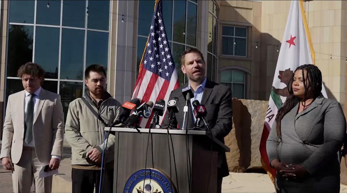 Eric Swalwell in a brown suit and open collar shirt stands at a podium and speaks into multiple microphones.