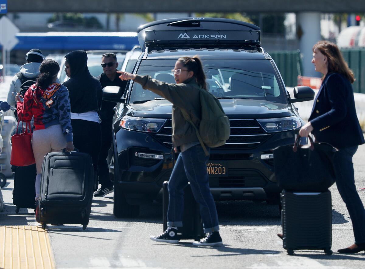 People walk with luggage in front of cars.