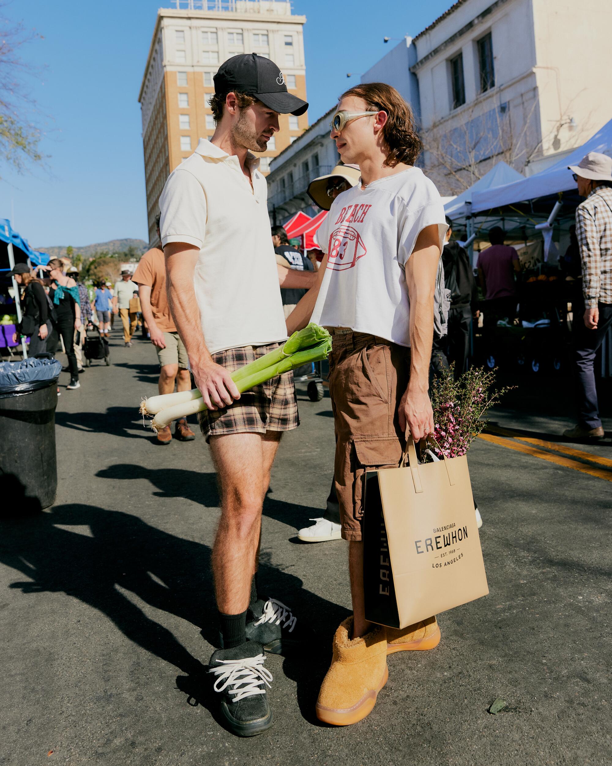 Austin wears a hat, polo top, shorts & sneakers. Carlos wears a top, shorts, boots and Balenciaga x Erewhon bag.