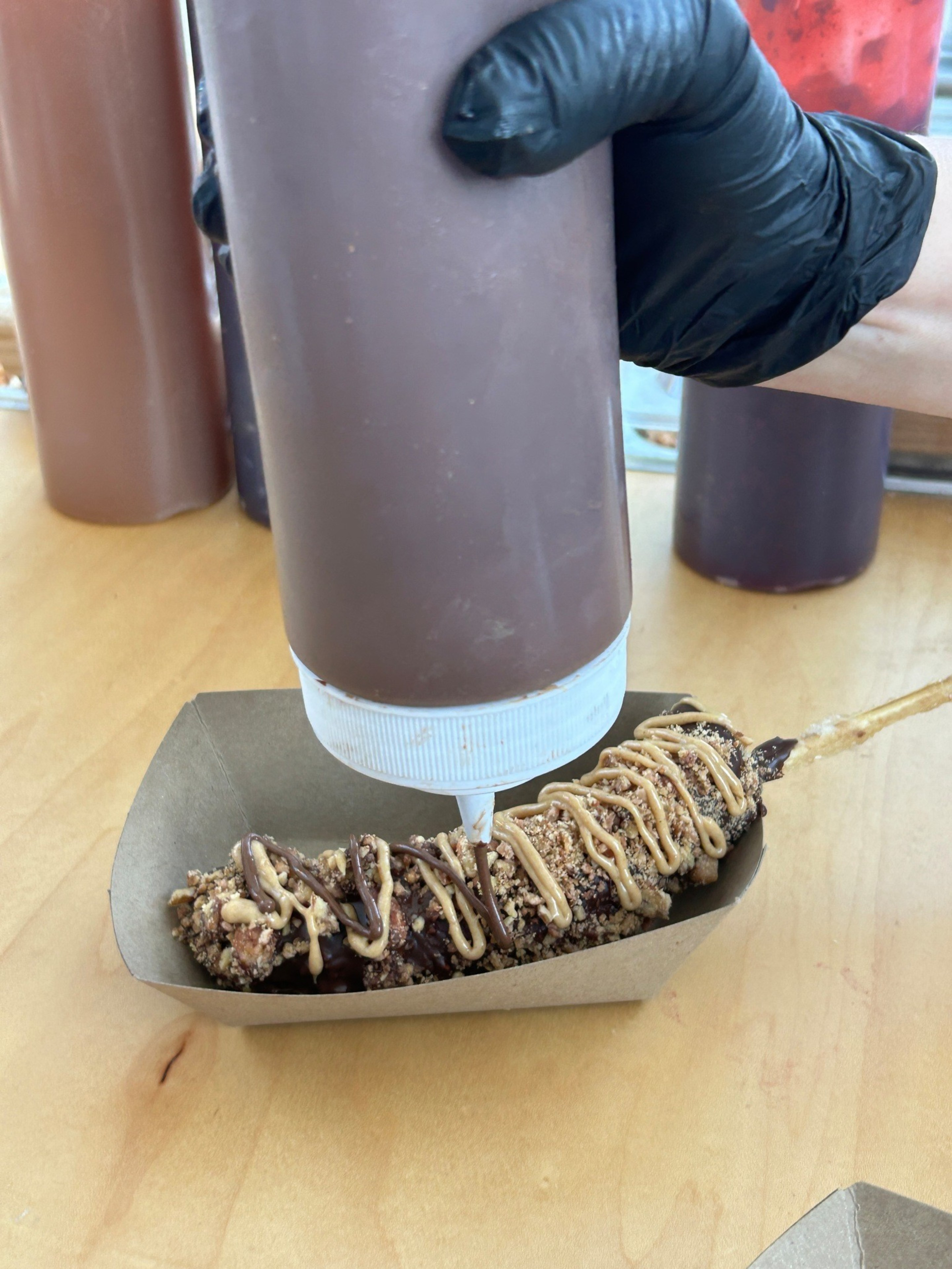 A gloved hand squeezes chocolate sauce from a bottle onto a stick coated with crushed nuts and drizzled with peanut butter in a paper tray.