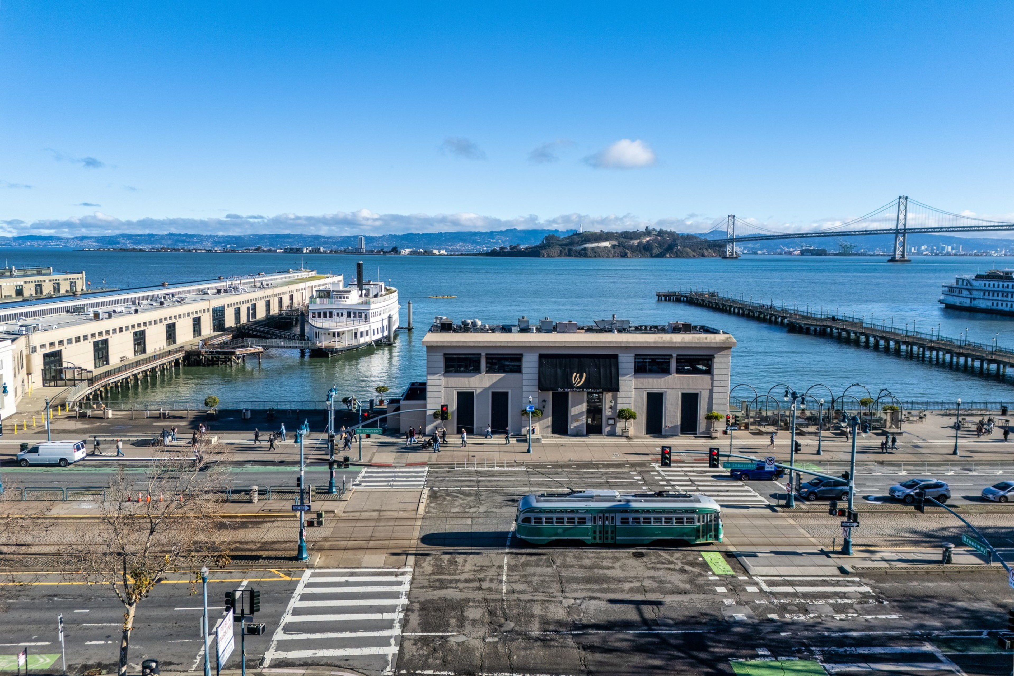 A waterfront scene includes a tram on a street with crosswalks, piers extending into the water, boats docked, a building with a black awning, and a distant bridge under a clear blue sky.