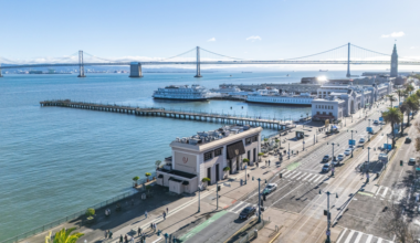 A waterfront city scene with piers, boats docked, a suspension bridge in the background, palm trees, and a busy street with cars and pedestrians.