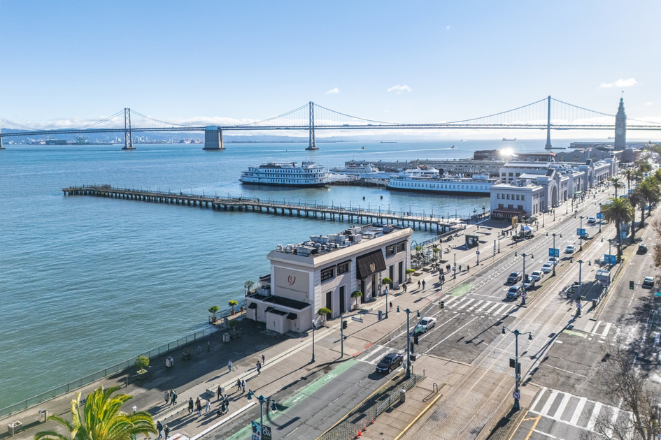 A waterfront city scene with piers, boats docked, a suspension bridge in the background, palm trees, and a busy street with cars and pedestrians.