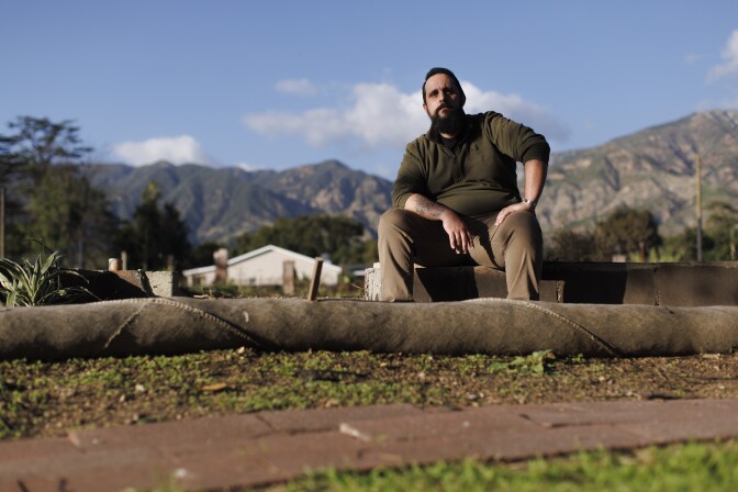 A man wearing brown sits on a cleared lot with mountains rising behind him.