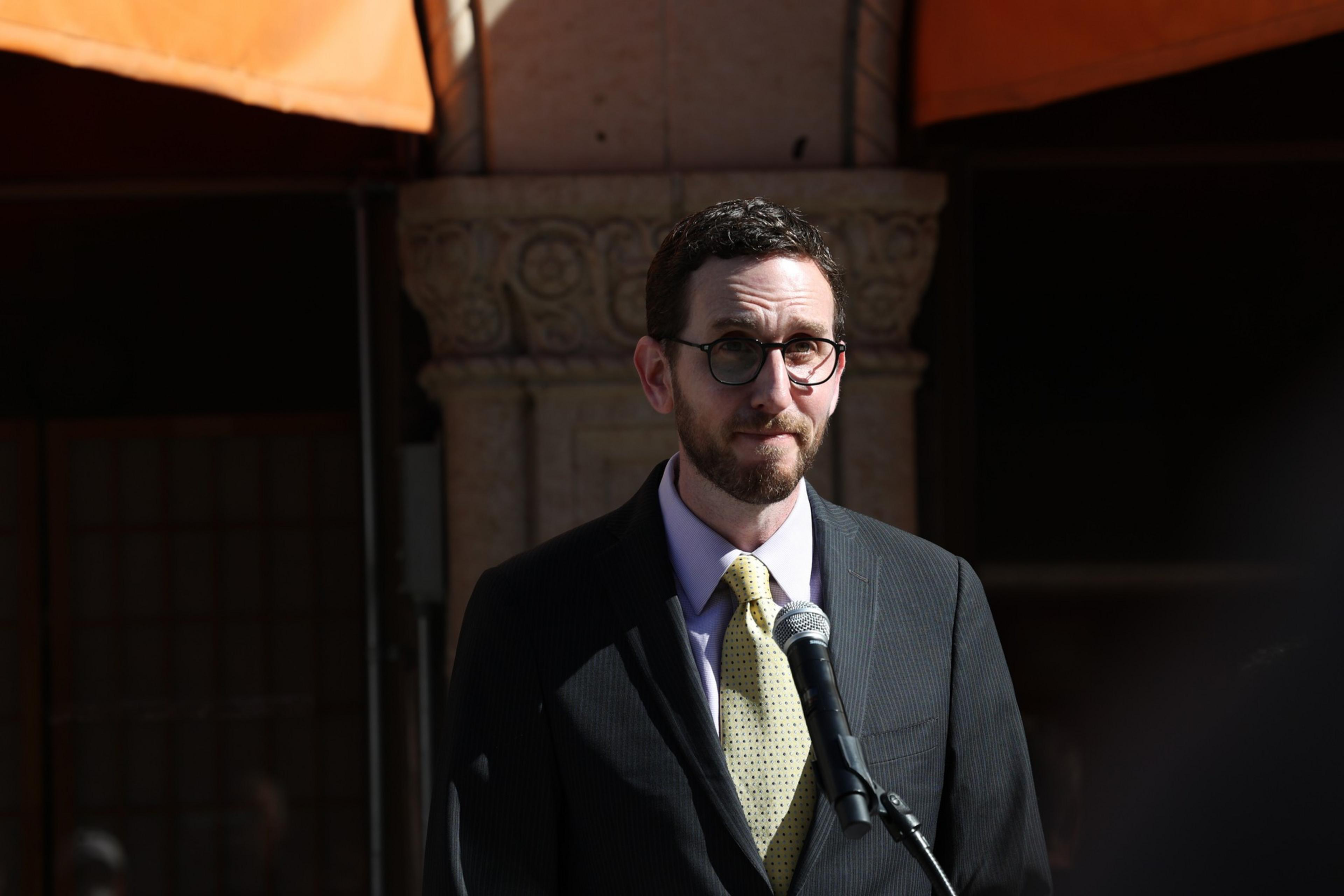 A man with glasses, a beard, and short curly hair wears a suit and yellow tie, standing behind a microphone, with ornate stonework and awnings behind him.