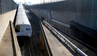 A white BART train approaches in a sunken concrete track area lined with fencing topped with barbed wire on both sides.