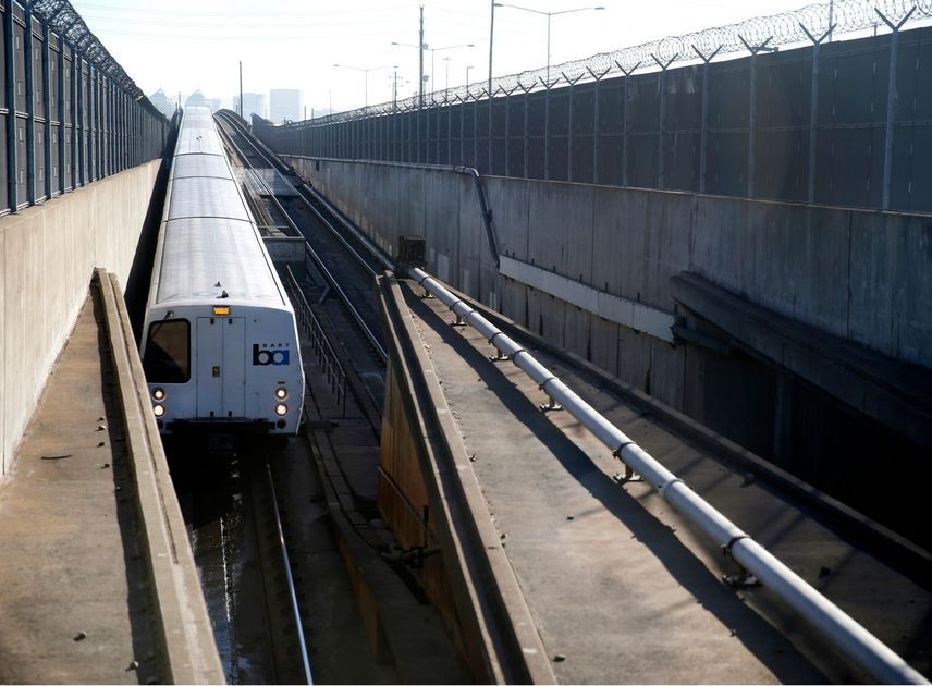 A white BART train approaches in a sunken concrete track area lined with fencing topped with barbed wire on both sides.