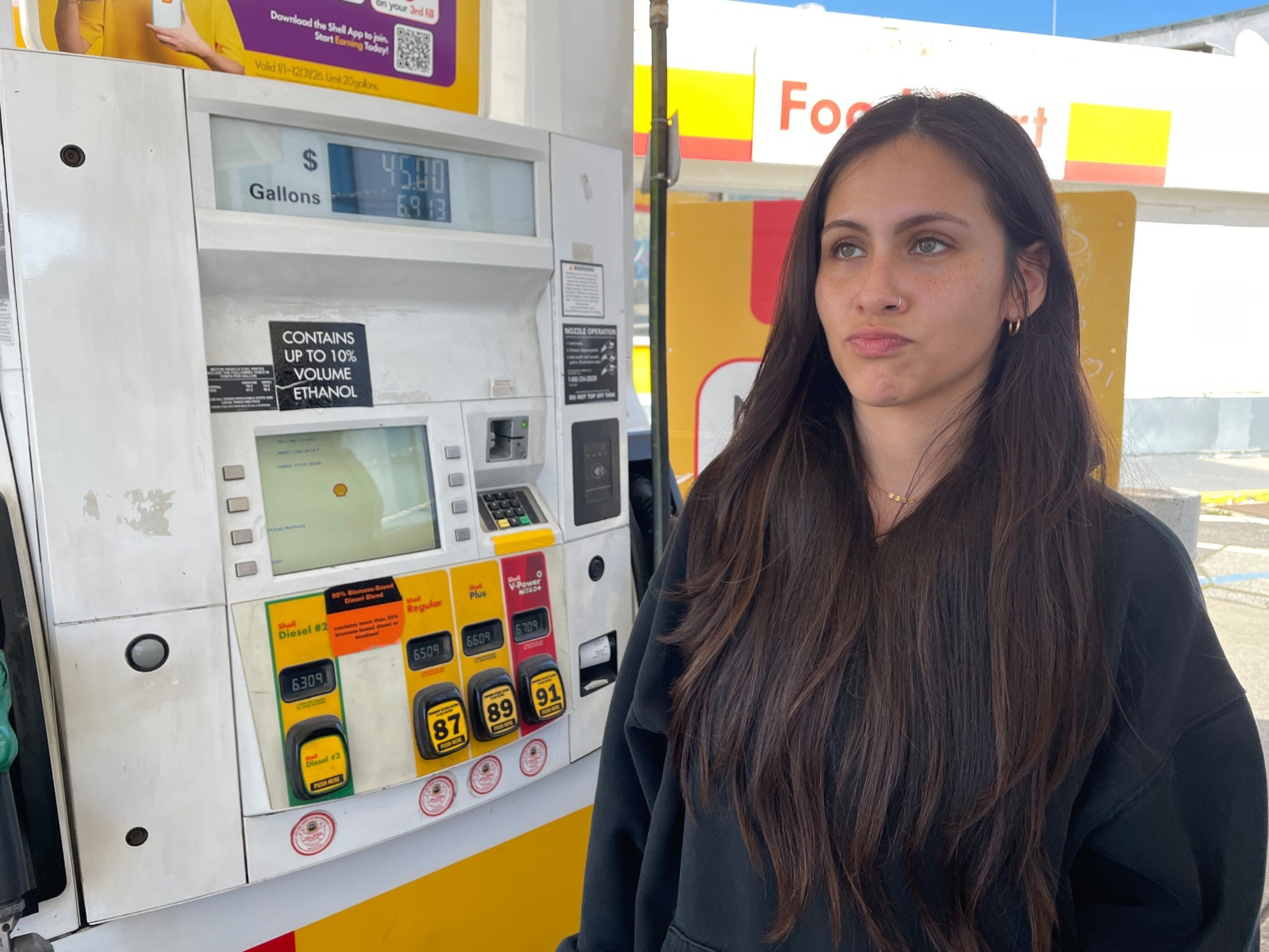 A woman with long dark hair and a black hoodie stands at a gas pump displaying $45.00 for 6.913 gallons at a Shell station.