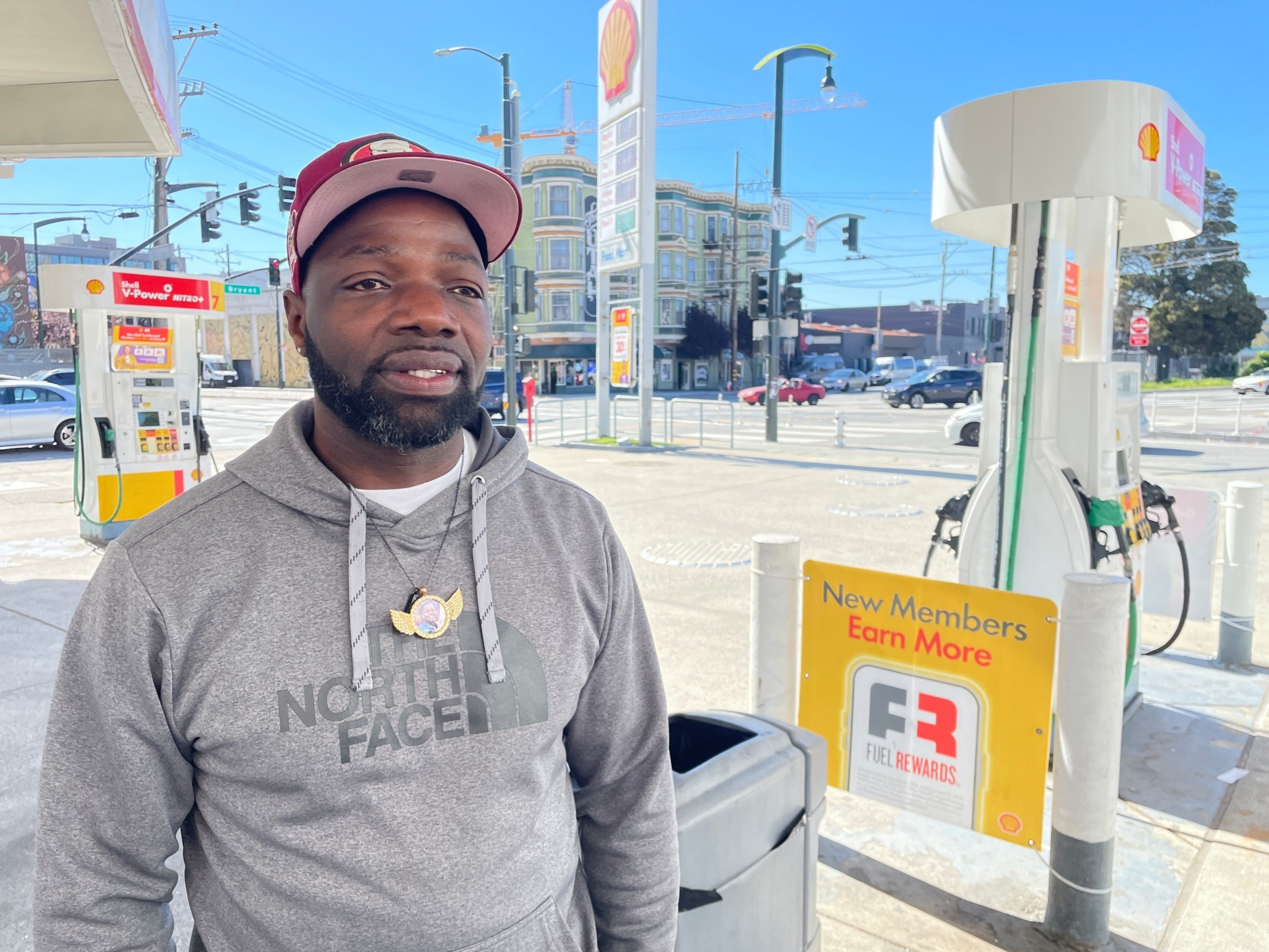 A man wearing a gray North Face hoodie and a red baseball cap stands at a Shell gas station near fuel pumps with a “New Members Earn More” Fuel Rewards sign.