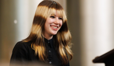 A crowd of young people, many holding phones, with one person holding a colorful sign that says “Congrats Ilyssa Liu, You’re Cooler Than Ice.”