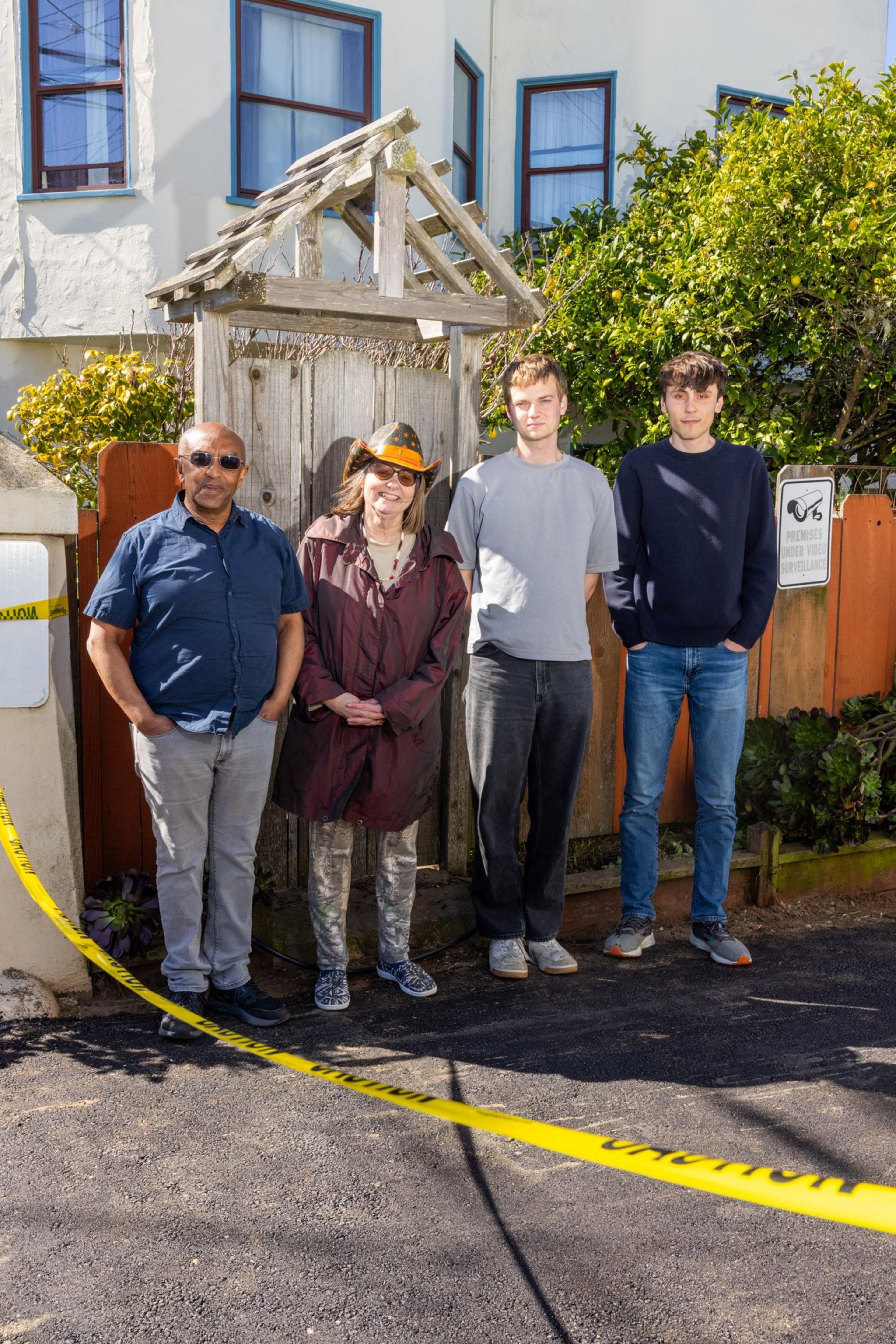Four people stand side by side on a paved path near a wooden gate with greenery behind them, with yellow caution tape stretched across the foreground.