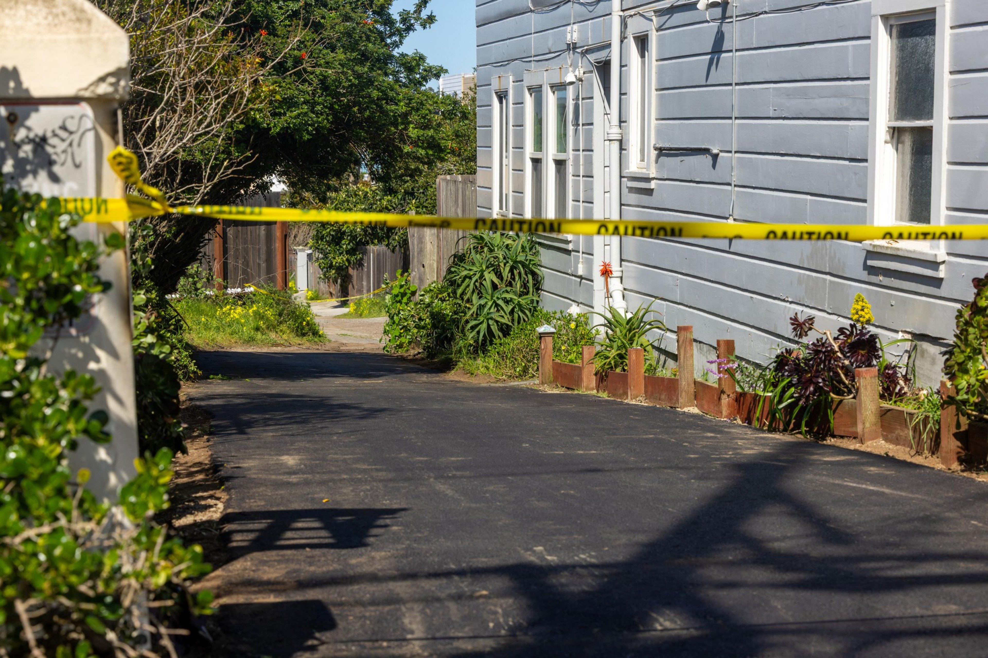 A narrow paved driveway beside a gray house is blocked by yellow caution tape, with plants and bushes lining the sides.