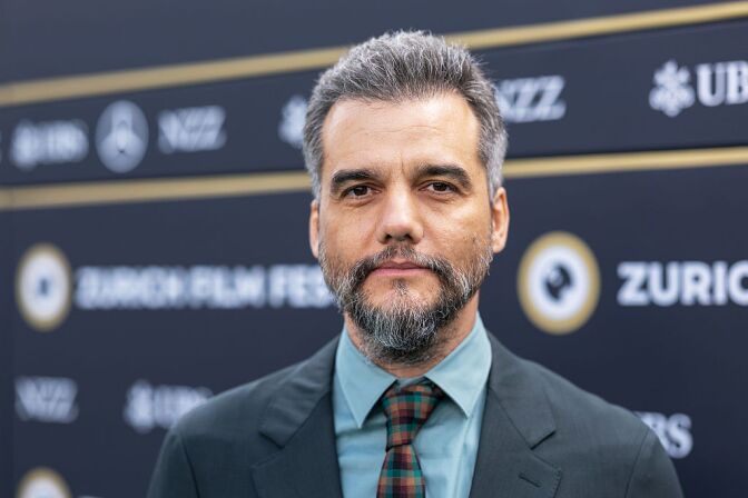A medium-light-skinned man with a beard and gray hair wears a suit and stares at the camera.