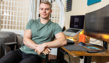 A man in a green shirt sits relaxed at a wooden desk with dual monitors, a laptop, and soundproofing panels on the wall behind him.