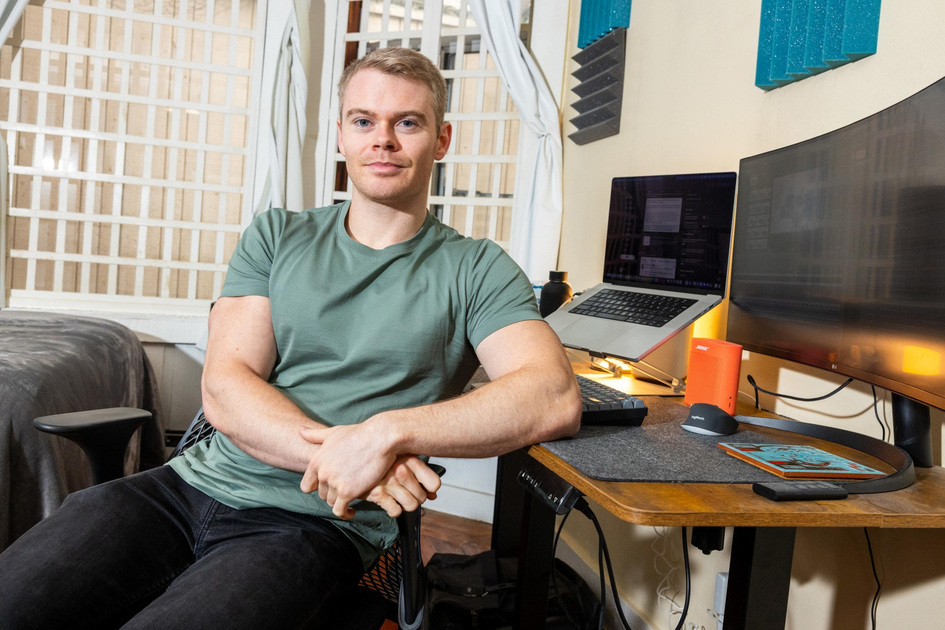 A man in a green shirt sits relaxed at a wooden desk with dual monitors, a laptop, and soundproofing panels on the wall behind him.
