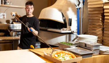 A man wearing a black shirt uses a long-handled peel to take a pizza out of a stone oven, with a stack of pizza boxes and restaurant supplies nearby.