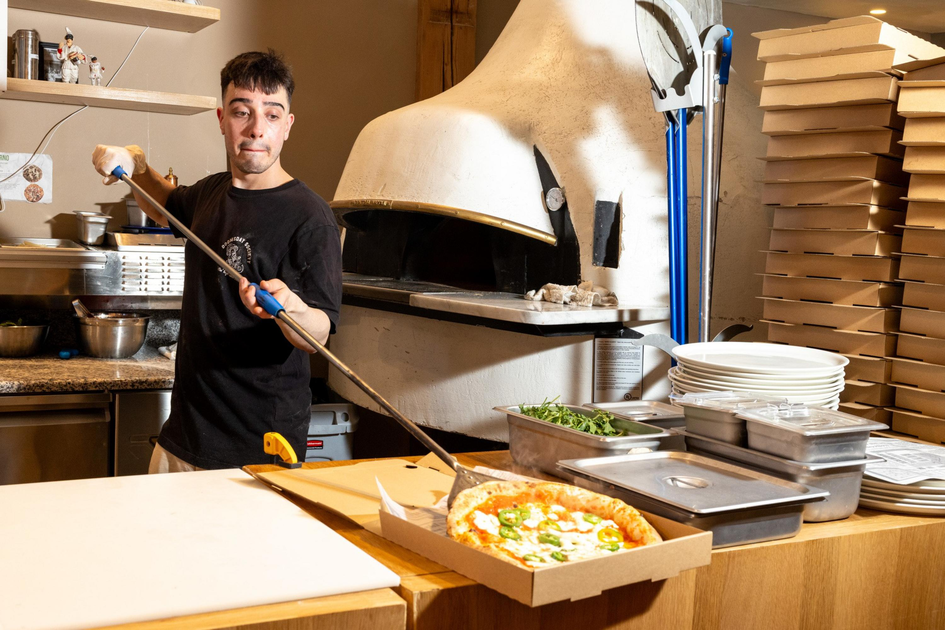A man wearing a black shirt uses a long-handled peel to take a pizza out of a stone oven, with a stack of pizza boxes and restaurant supplies nearby.